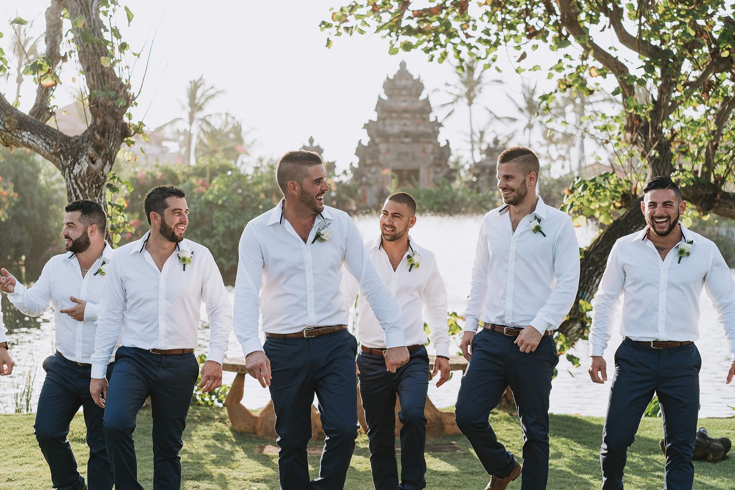 Groomsmen in white shirts and navy pants pose together in a tropical garden setting.