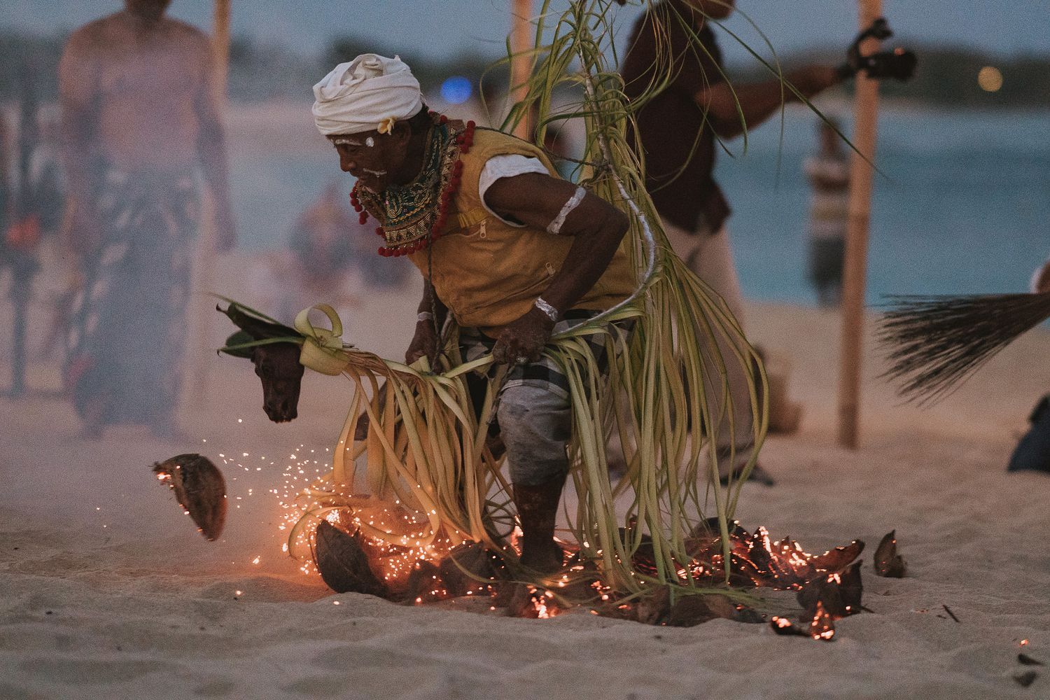 A traditional fire dancer performs on a sandy beach at dusk wearing grass skirt and ceremonial attire.
