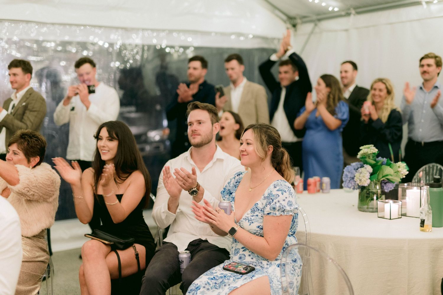 Wedding guests in formal attire applaud during reception celebration at decorated indoor venue.