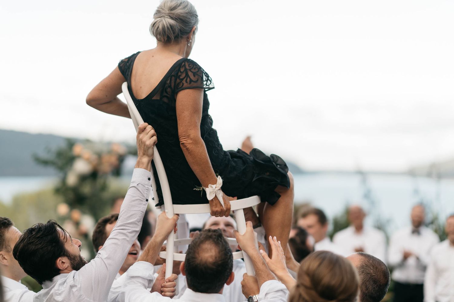 People in white outfits lifting someone in a black dress during outdoor celebration with water in background.