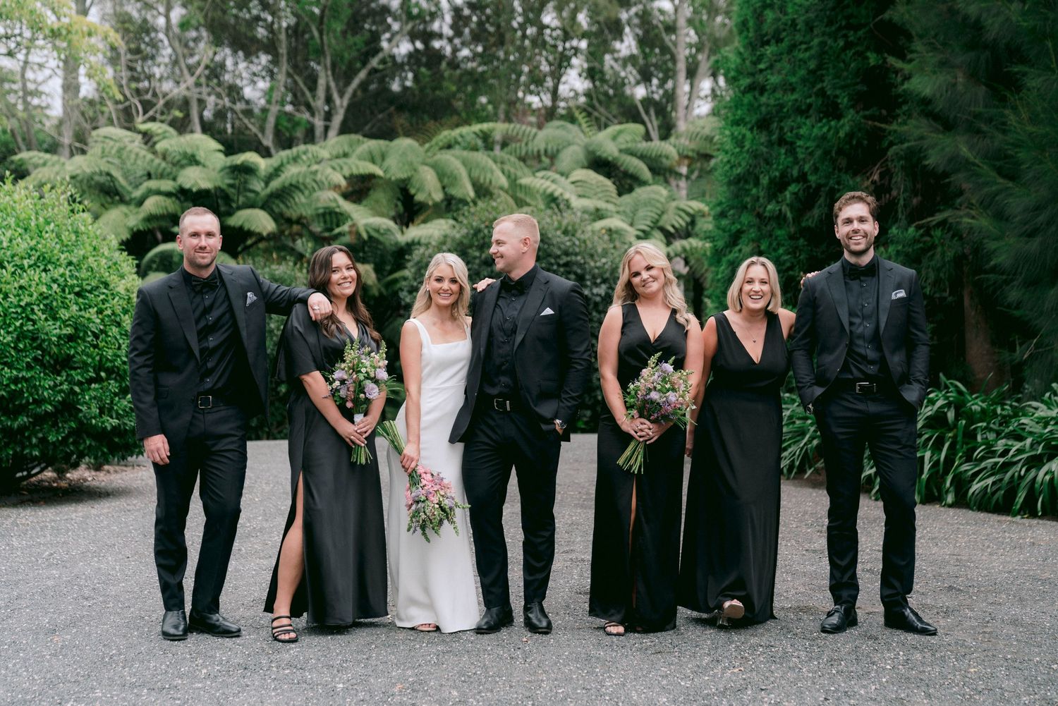A wedding party in black and white formal attire poses together on a garden path.