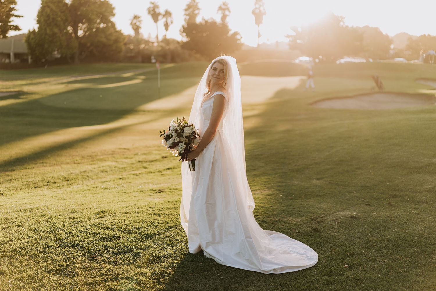 A flowing white wedding dress moves gracefully across a sunlit golf course during golden hour.