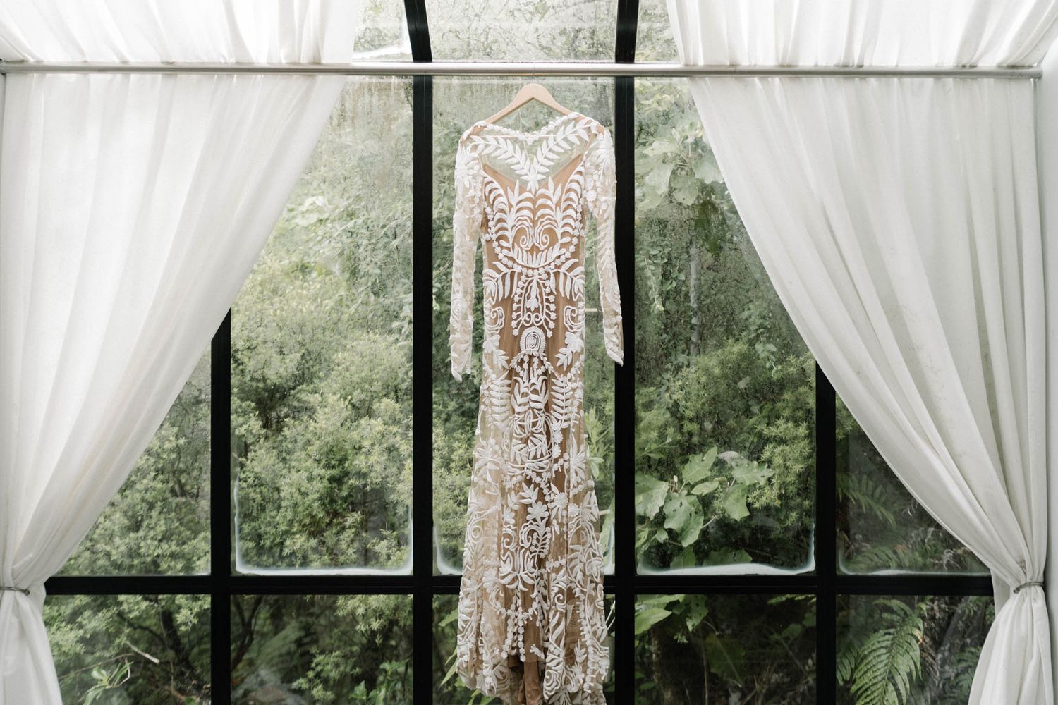A delicate white lace wedding dress hangs in front of large windows with flowing sheer curtains and green foliage outside.