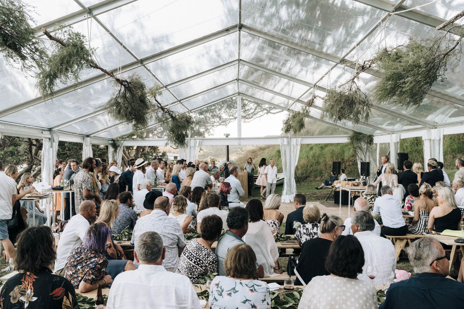 Elegant white tent reception venue decorated with greenery and filled with wedding guests celebrating at decorated tables.