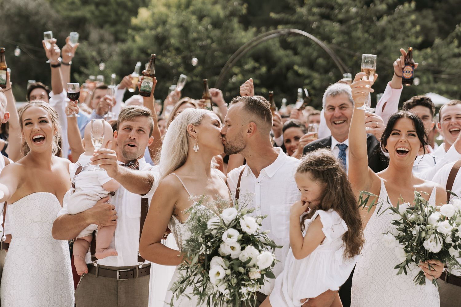 Wedding party and guests gather outdoors for celebratory photo holding white bouquets and smiling with joy.