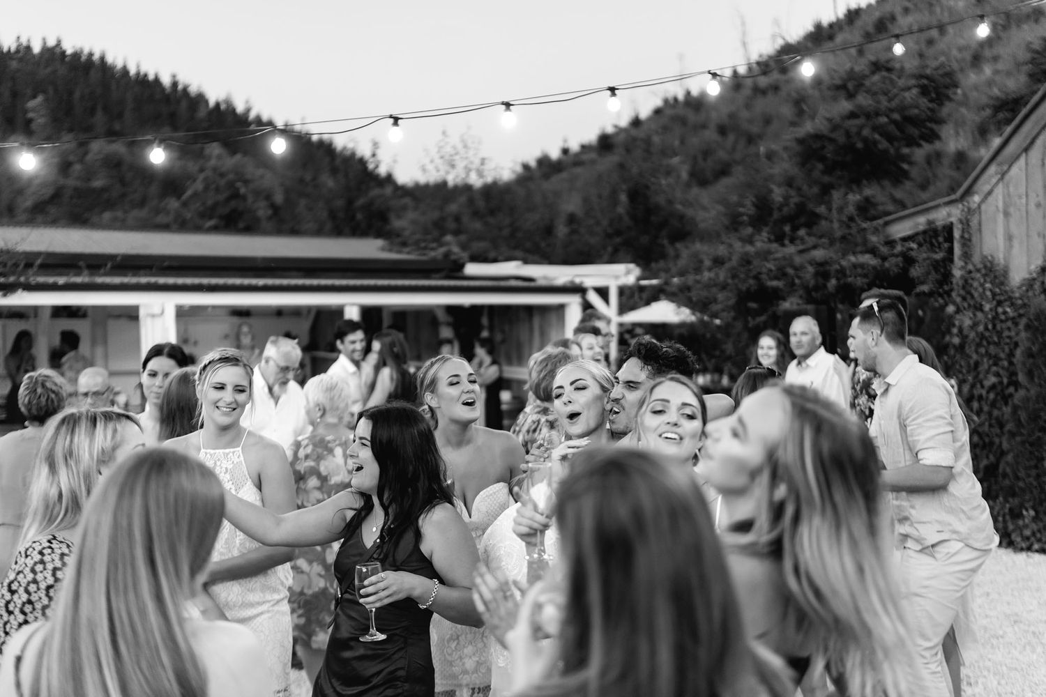 Black and white photo of guests dancing and celebrating at an outdoor party with string lights and mountain backdrop.