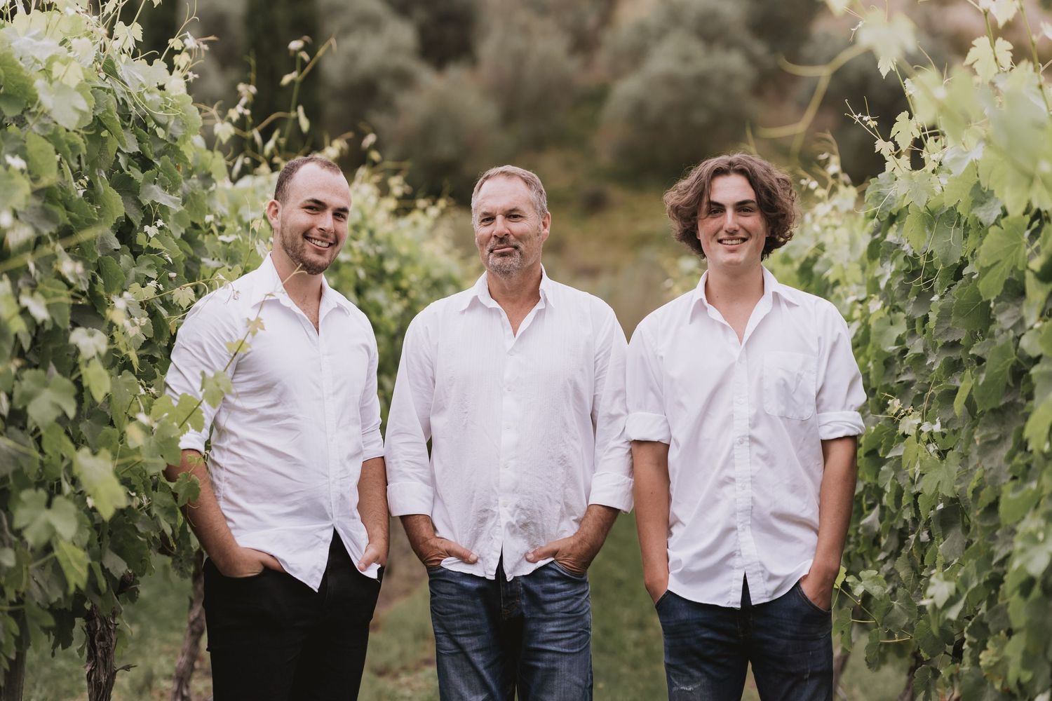 Three people in white shirts stand together smiling in a lush vineyard on a sunny afternoon.