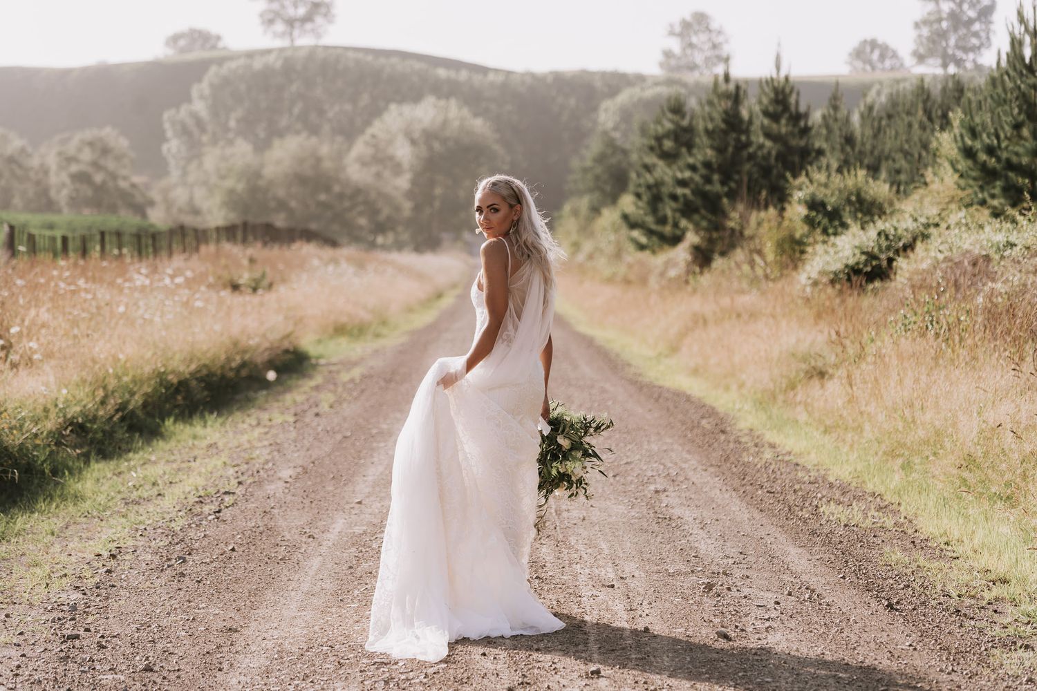 A bride in a flowing white dress stands on a rural dirt road surrounded by golden dry grass and evergreen trees.