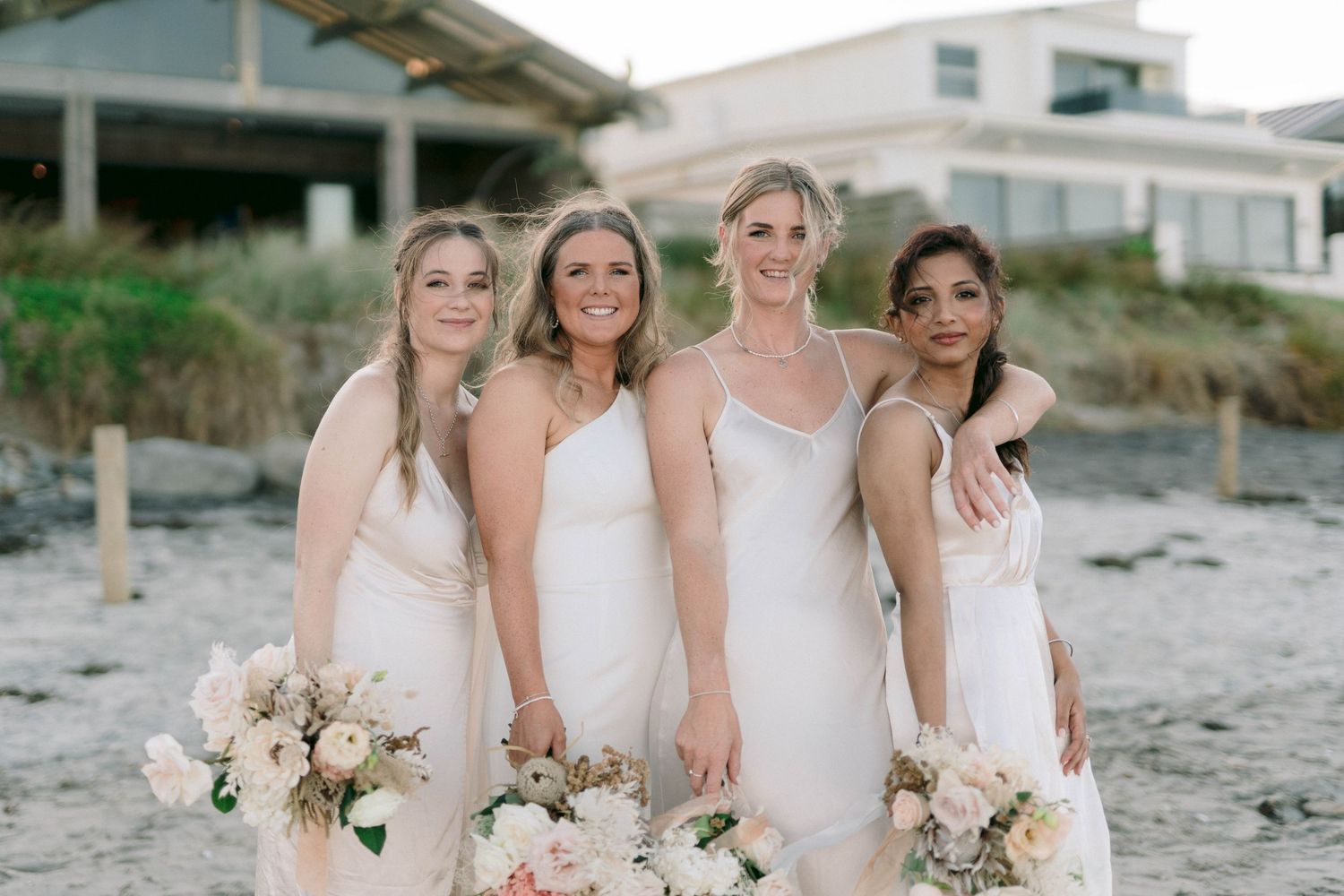 Bridesmaids in white dresses hold bouquets while standing on a sandy beach.