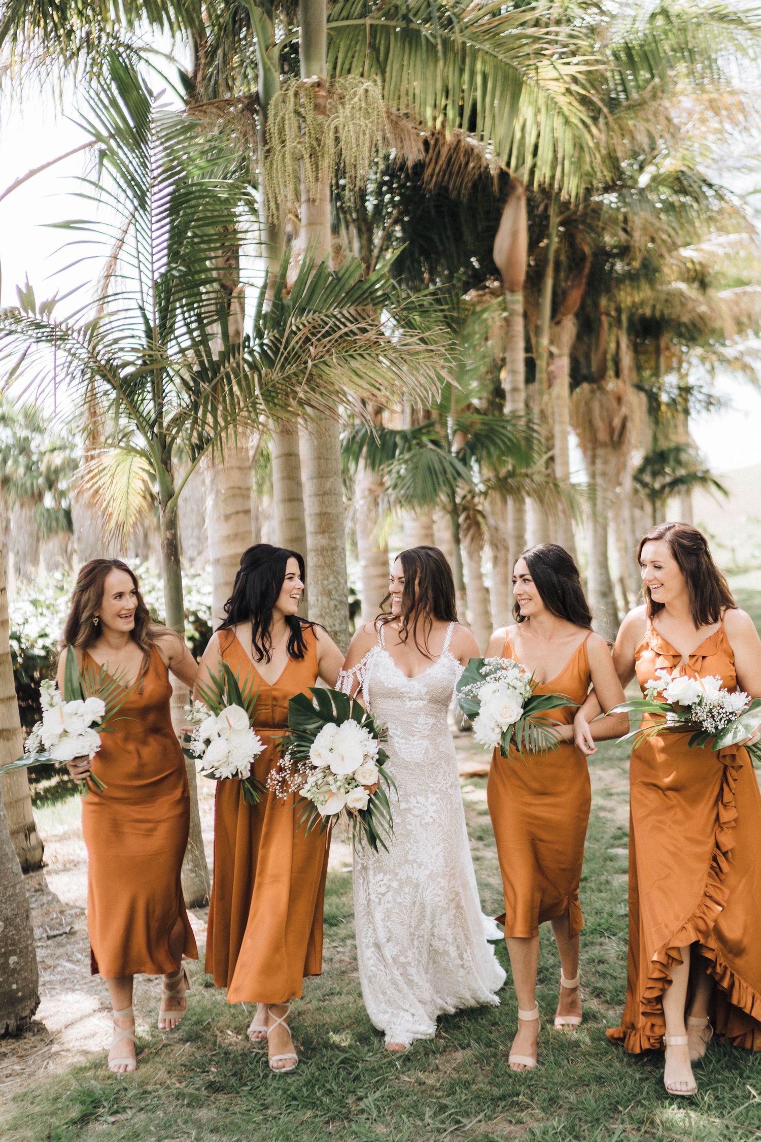 Wedding party in orange bridesmaid dresses and white bridal gown posing among palm trees.