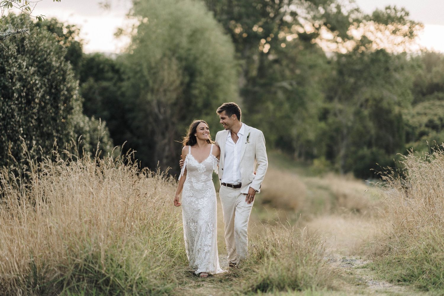 A couple in white attire walks together on a rural path surrounded by tall golden grass at sunset.
