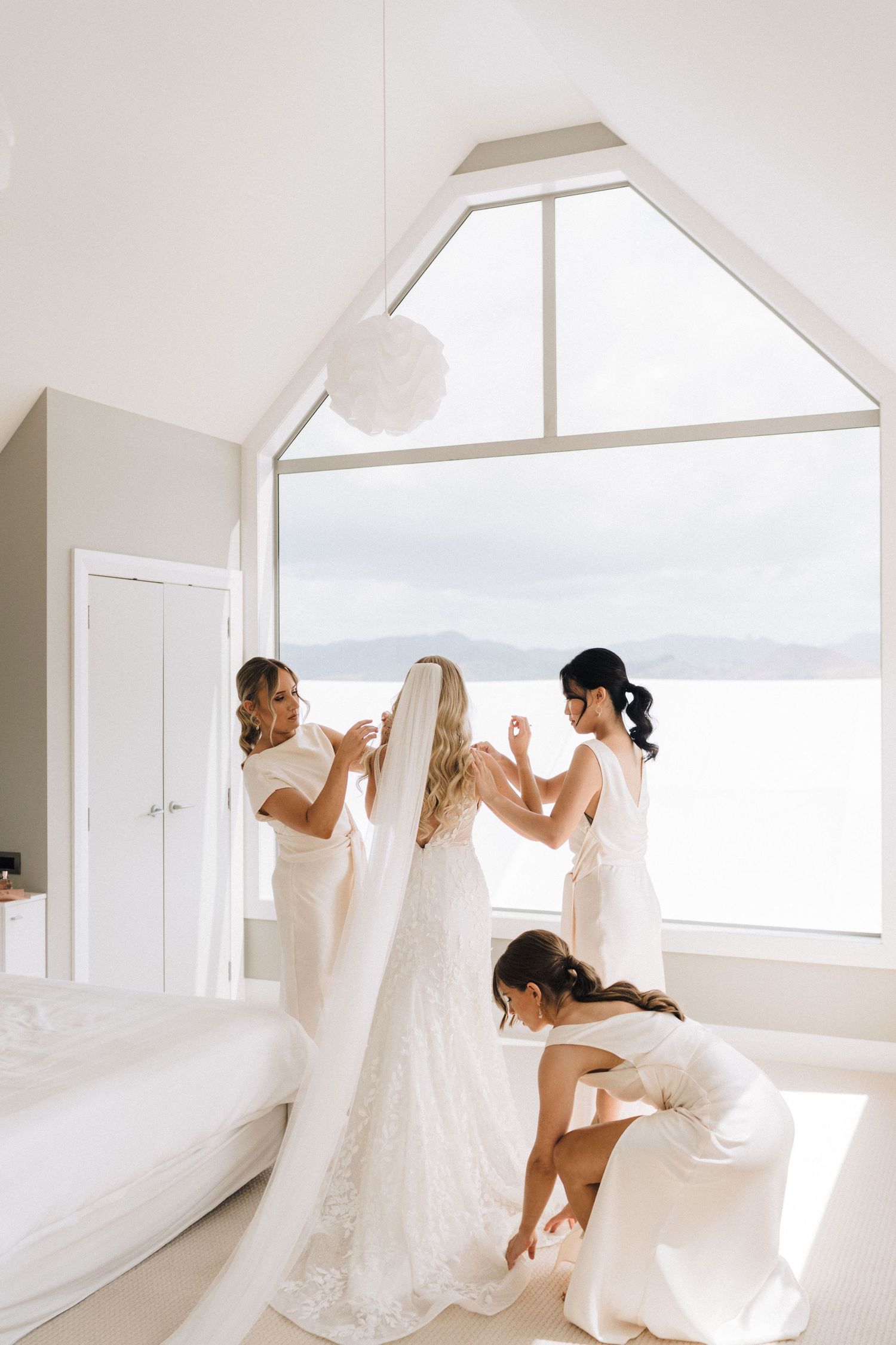 Bridesmaids helping bride get ready in bright room with panoramic mountain view.