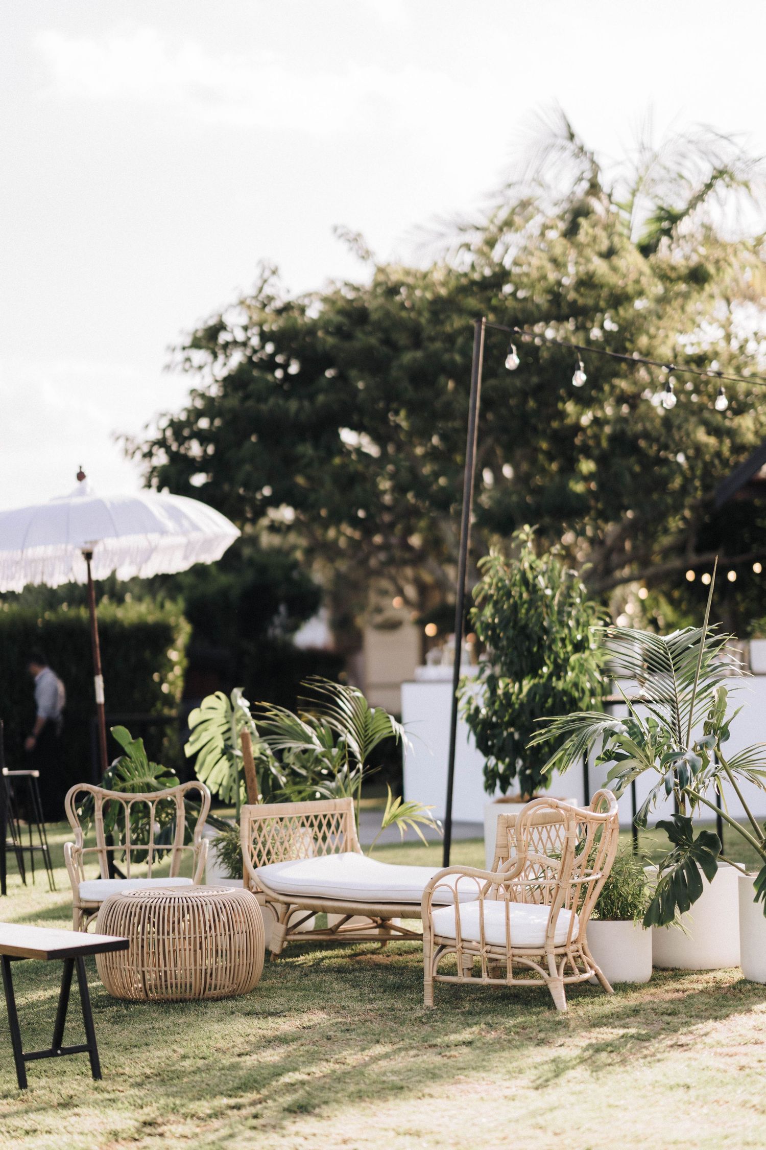 Outdoor lounge area with rattan furniture and white parasols set up on a lawn with trees.