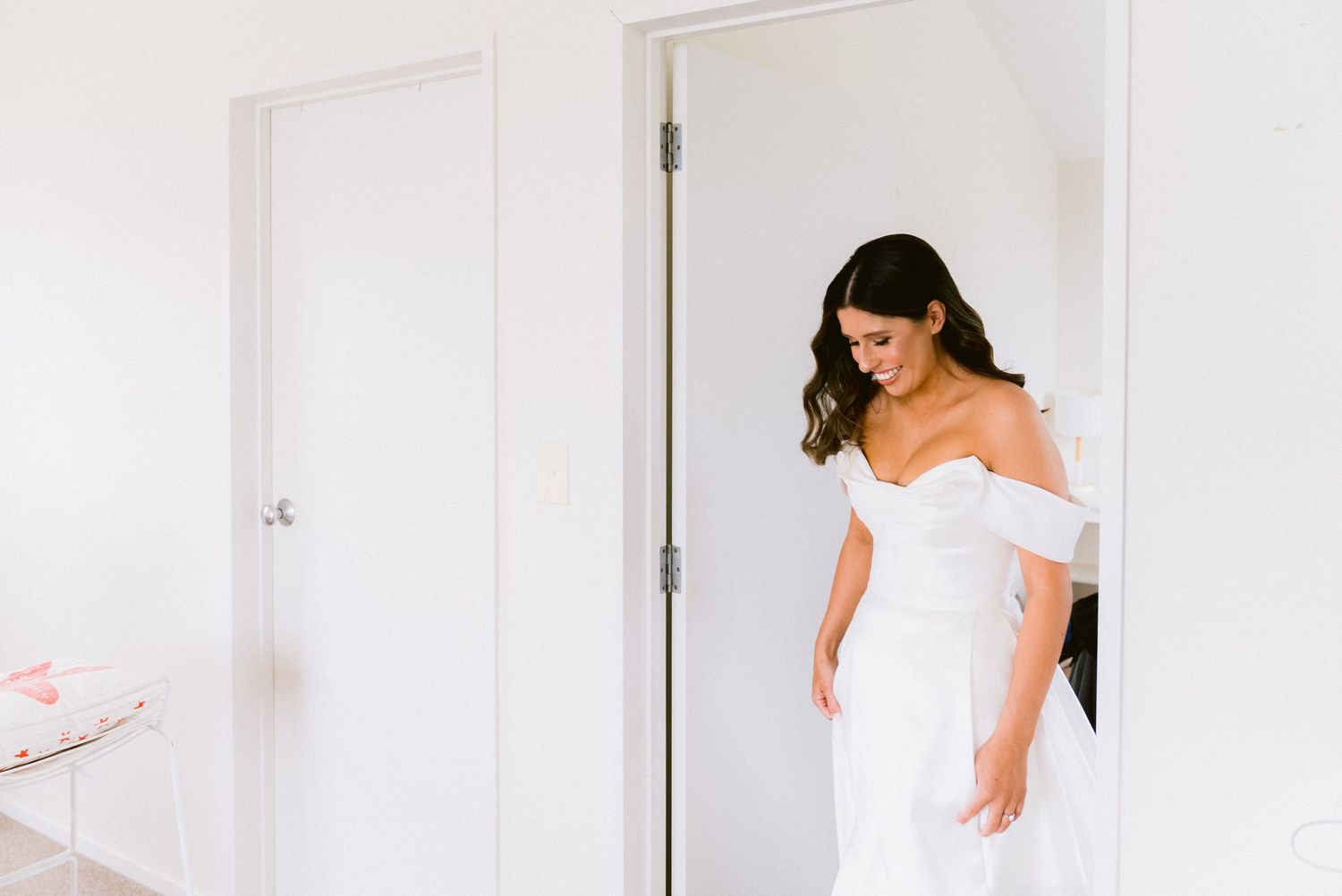 Someone in an elegant off-shoulder white wedding dress stands in a bright room looking down at their gown.