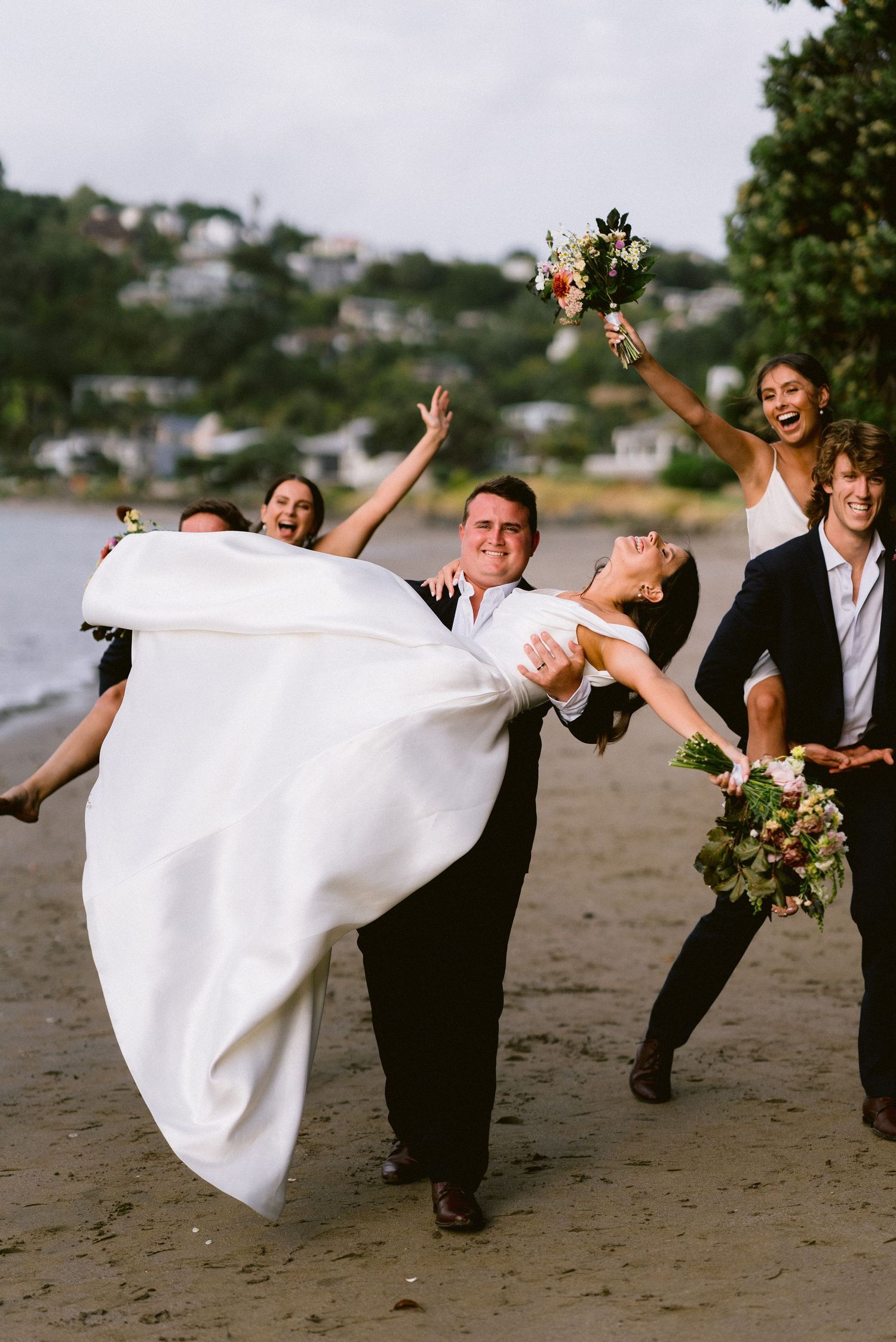 Wedding party performs playful lifting poses on a beach at sunset.