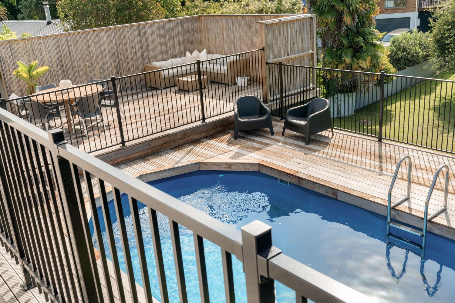 Above-ground swimming pool with black safety fence on wooden deck in backyard.