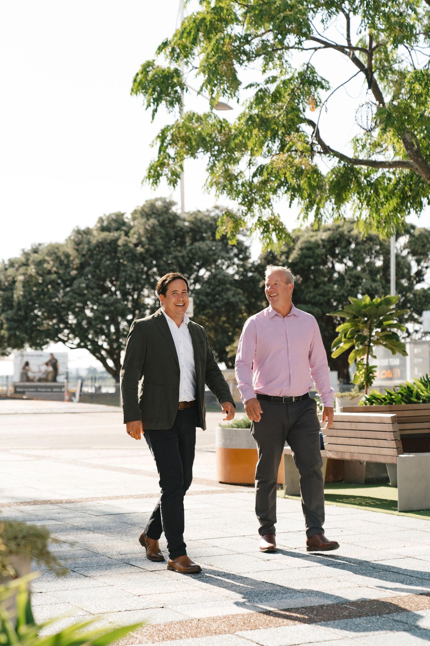 Two professionals walking together on a path with trees and benches in the background.