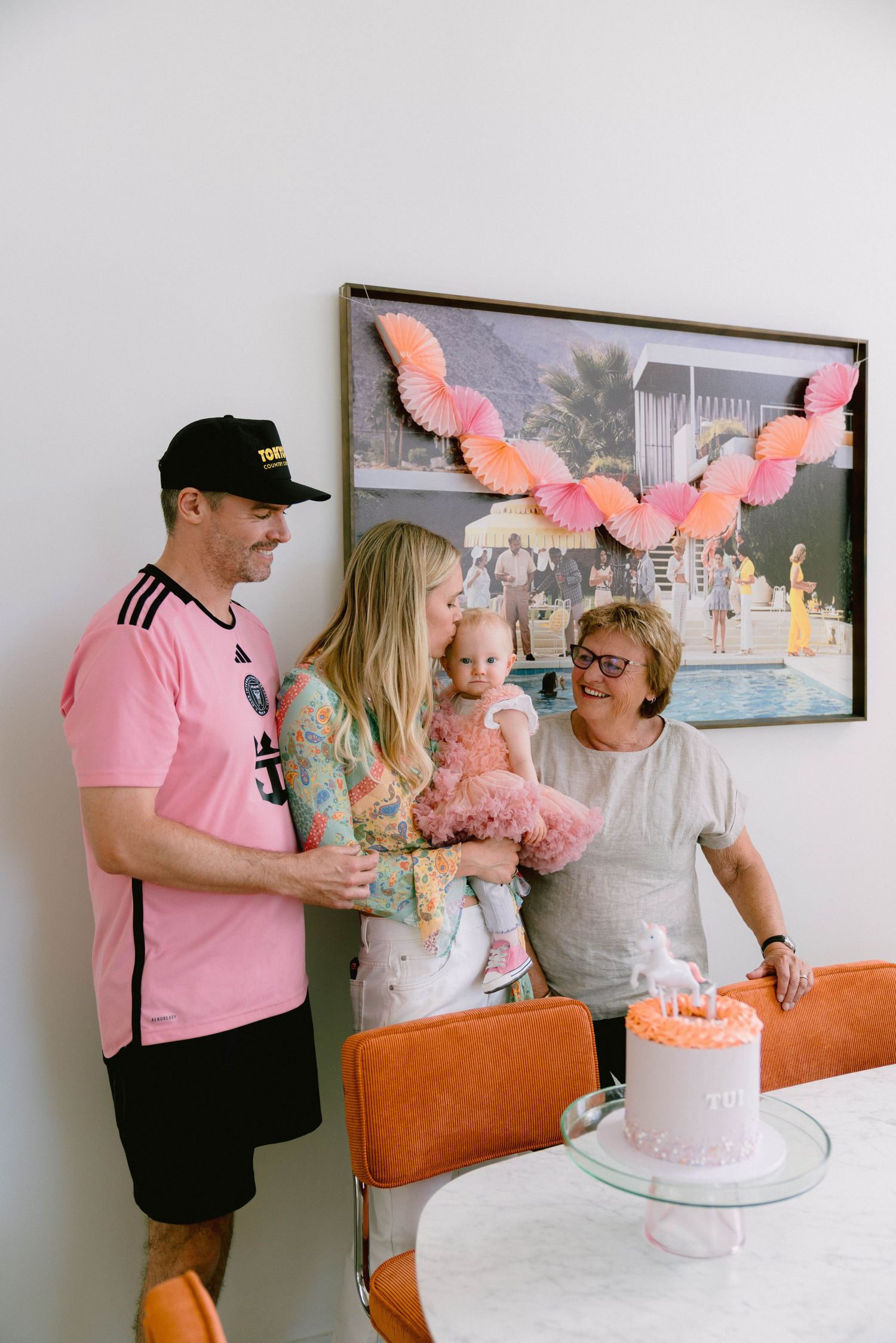 A small party gathering around a pink frosted birthday cake on a white table in a modern interior setting.