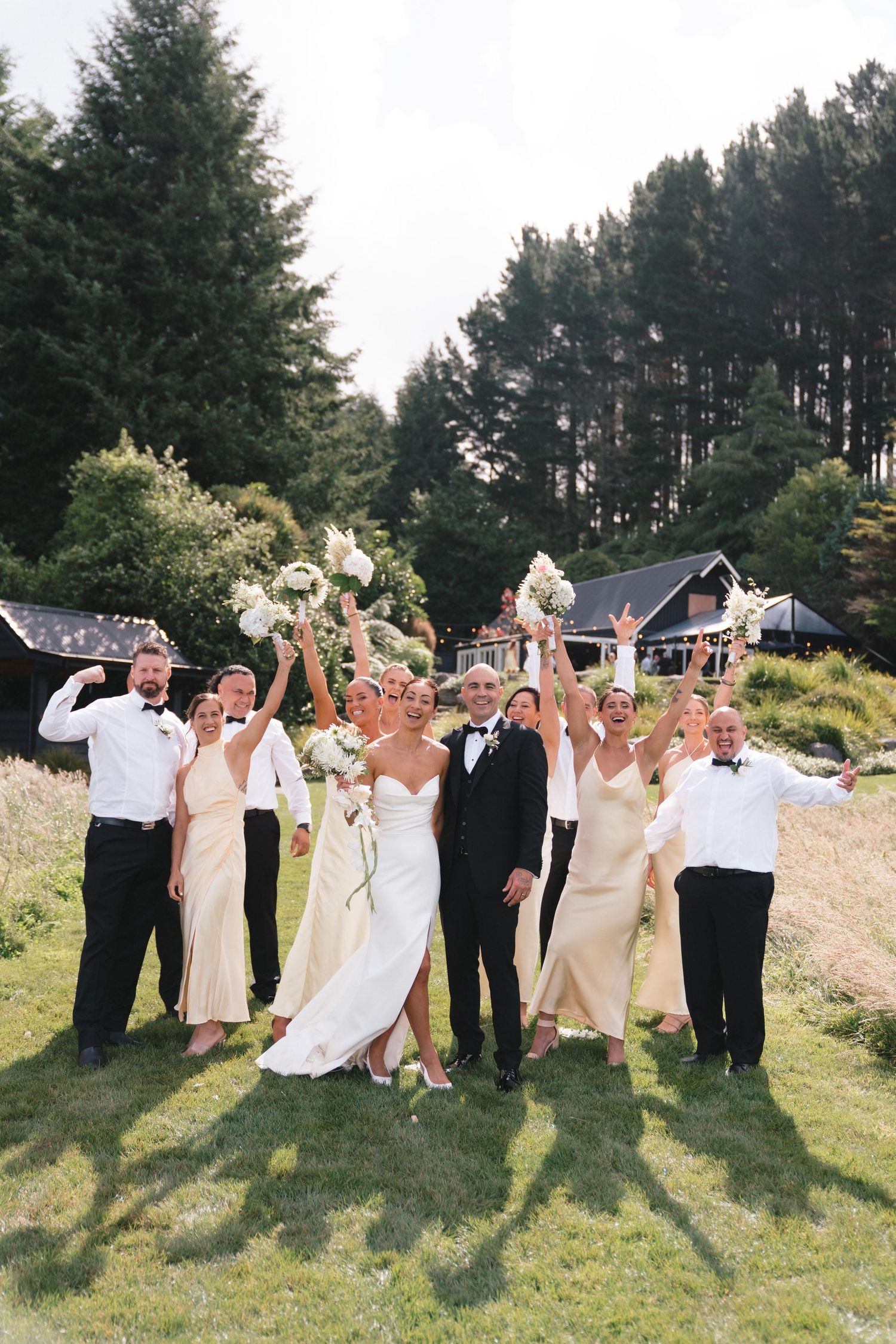 Celebratory wedding group jumps and poses together on lawn with pine forest backdrop.