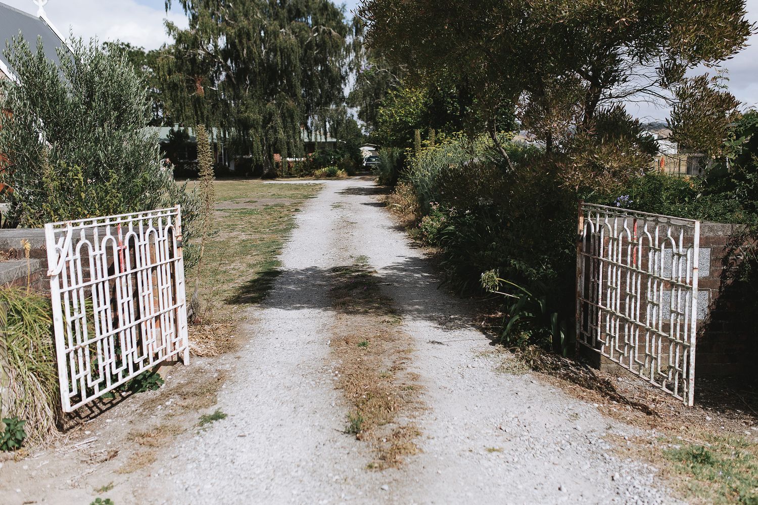 Rustic white metal gate opens onto a winding gravel path through a serene country garden setting.