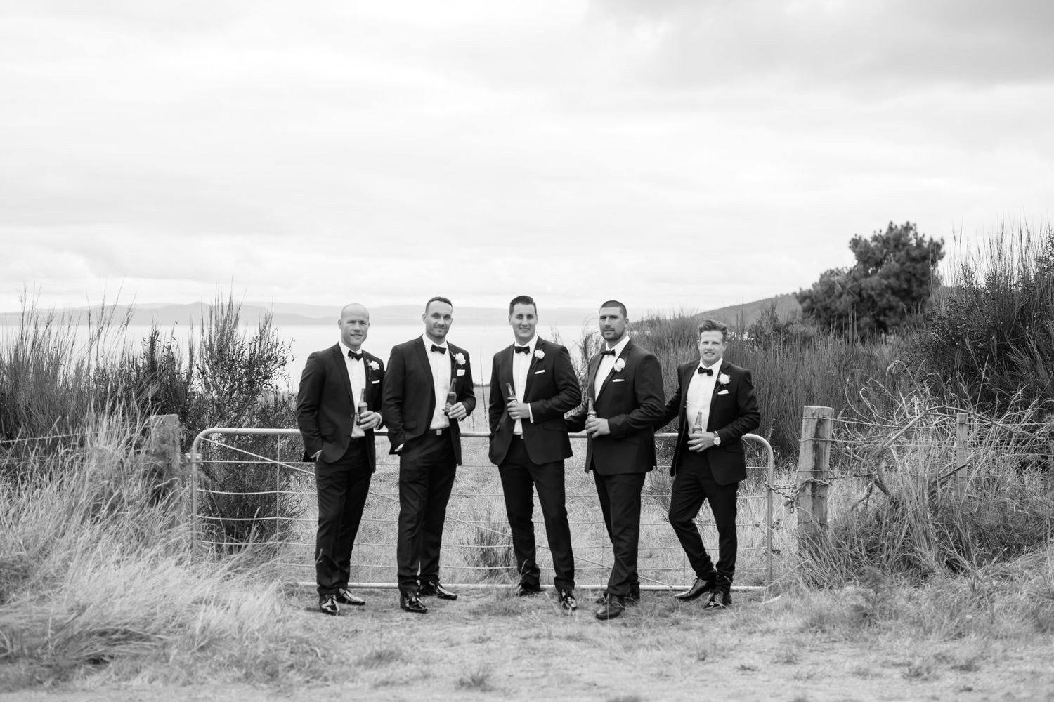 Wedding party stands in a dramatic black and white panoramic beach scene with tall grass creating natural framing.