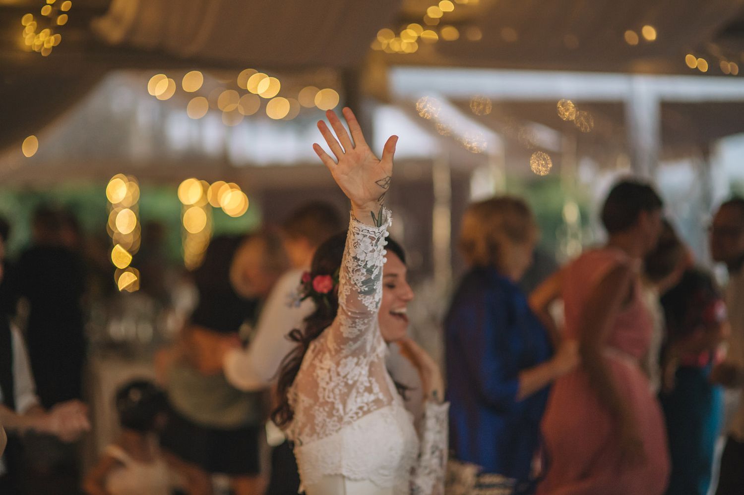 Wedding guests dance and celebrate on a dimly lit dance floor with glowing bokeh lights in the background.