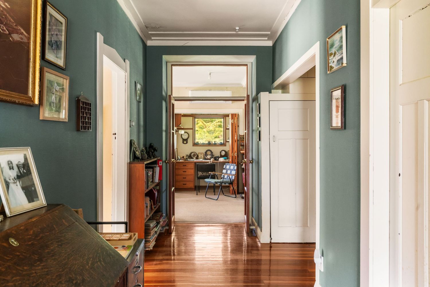 Long hallway with teal green walls, wooden floors and framed artwork leading to bright room.