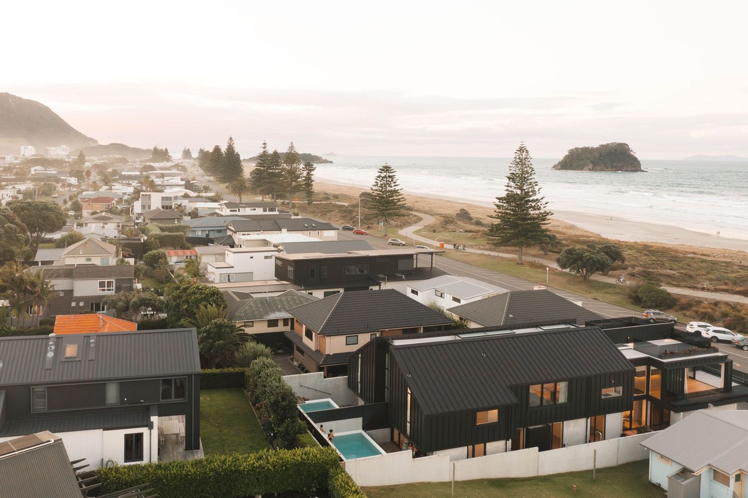 Aerial view of modern beachfront homes with dark roofs overlooking a misty coastal bay and sandy beach.
