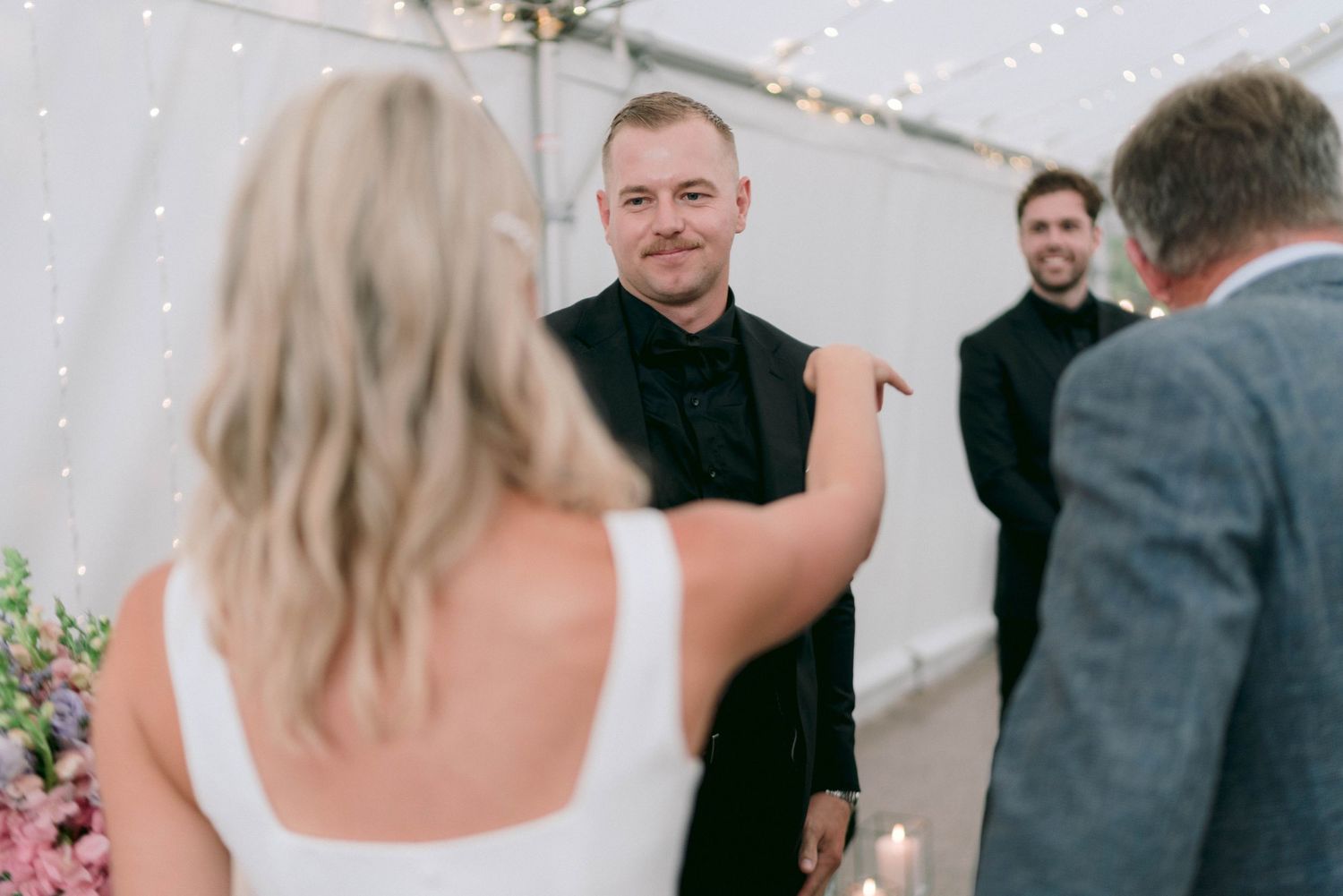 A wedding ceremony takes place under a white tent with string lights overhead.