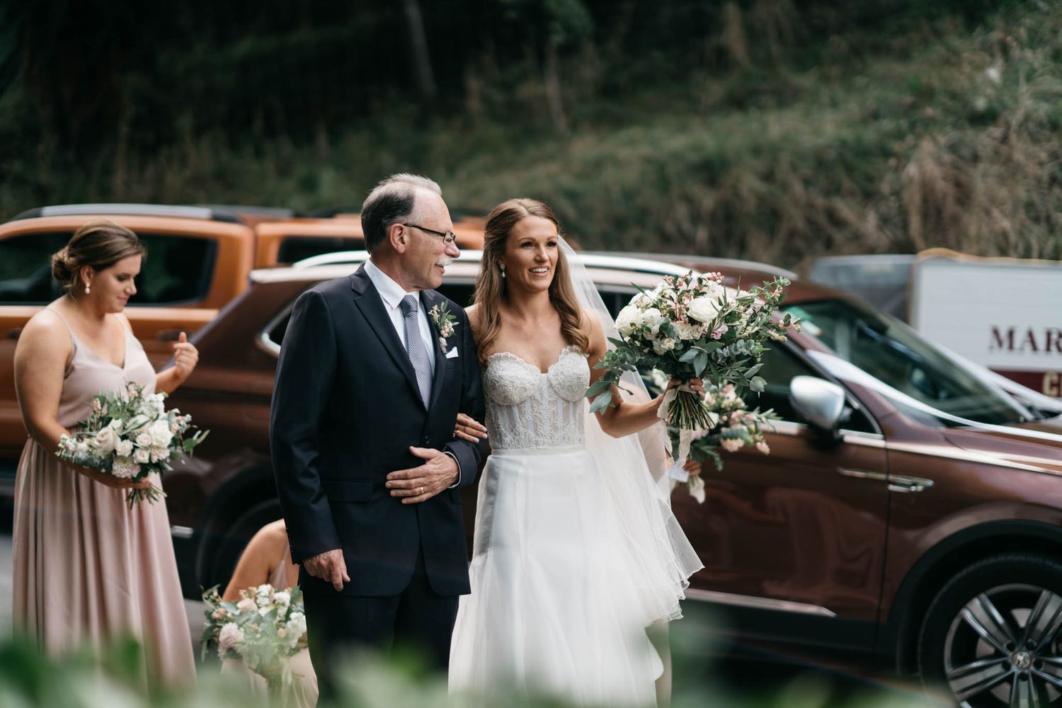 Joyful wedding party walks together outdoors near vintage cars while holding elegant white and green bouquets.