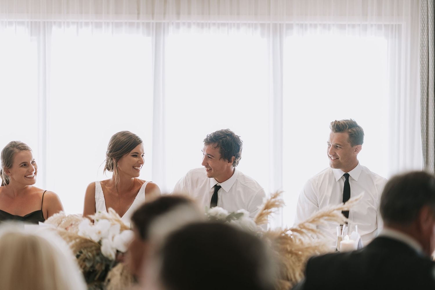 Lively wedding party members share laughs while seated at head table during reception celebration.