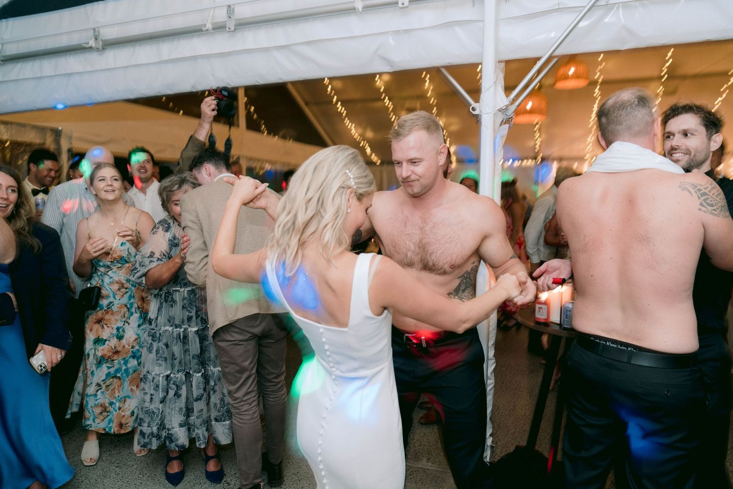 Wedding guests dancing and celebrating under a white tent during an evening reception party.