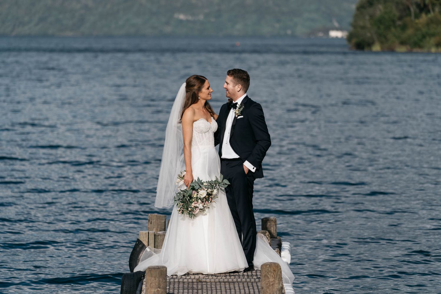 A couple stands together on a wooden dock by a serene lake with mountains in the background.