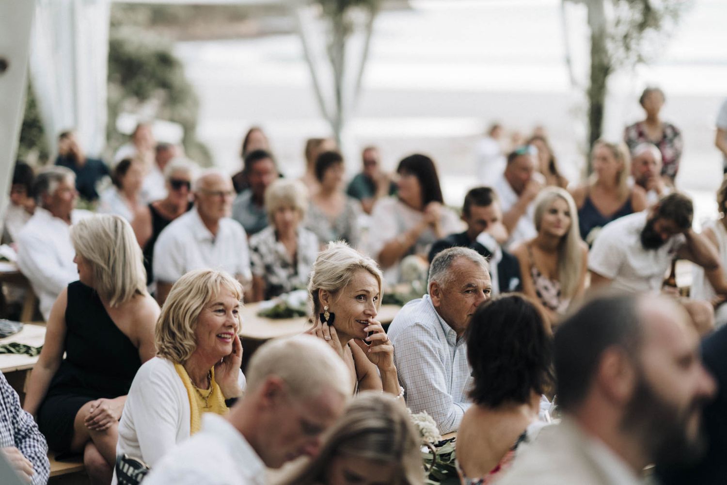 Wedding guests seated at tables smile and laugh during an outdoor reception celebration.