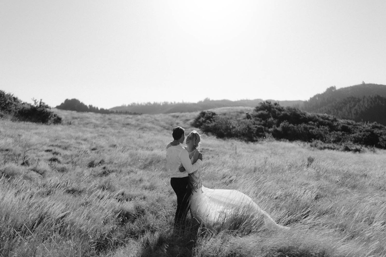 Two figures standing together in a windswept grassy meadow on a misty hillside captured in black and white.
