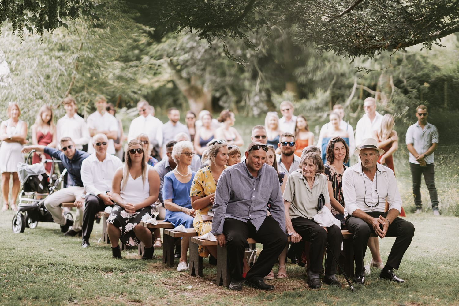 Wedding guests seated outdoors on white chairs during a summer garden ceremony, dressed in light summer attire.