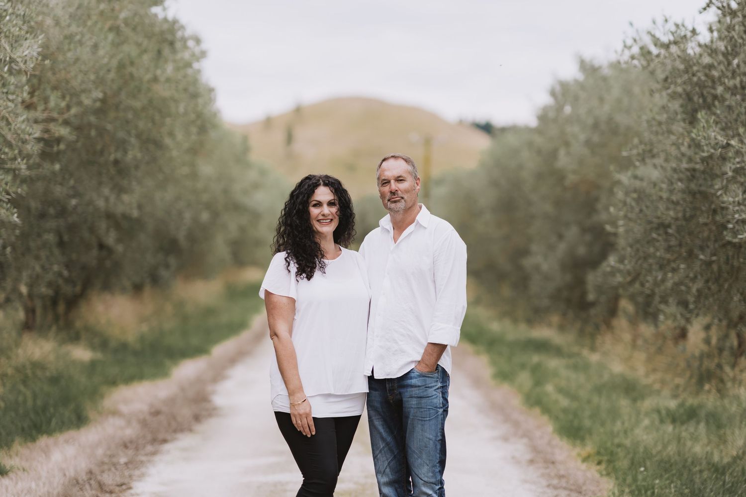 A couple walks down a scenic dirt path lined with olive trees on a sunny afternoon.