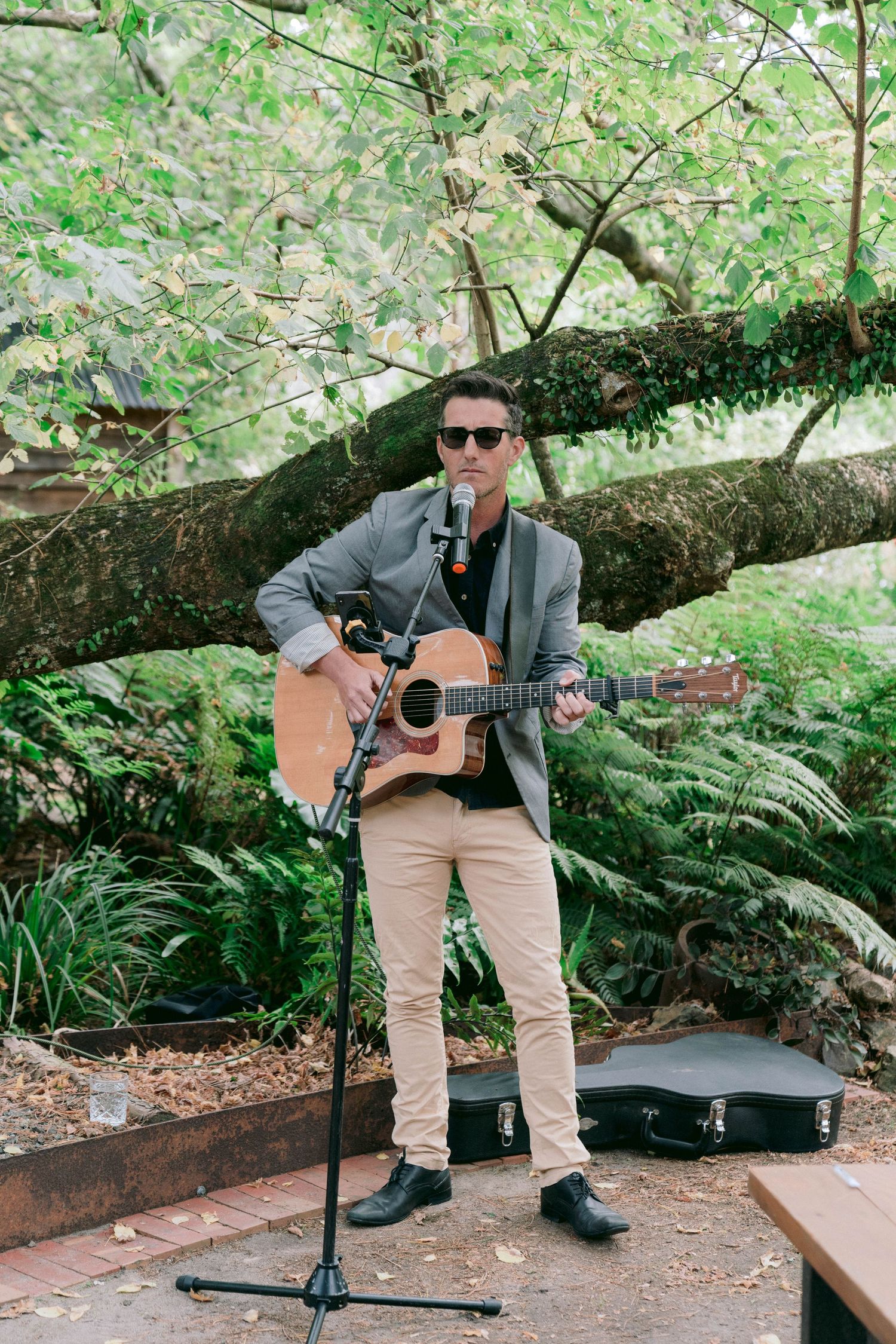 Musician in sunglasses plays acoustic guitar while seated on rustic outdoor bench surrounded by foliage.