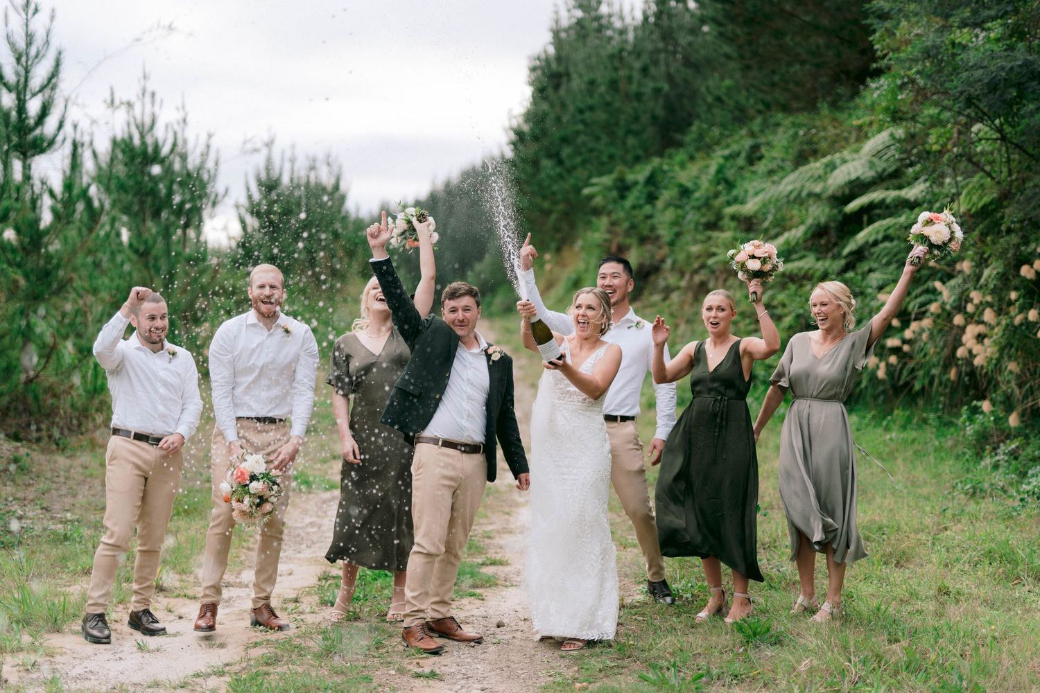 A wedding party celebrates outdoors while popping champagne bottles on a dirt path surrounded by greenery.