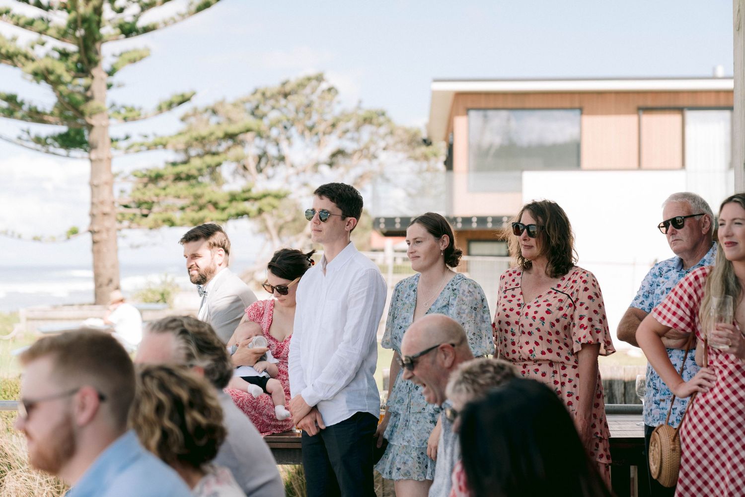 Outdoor wedding guests gather at a modern beachside venue on a sunny day.