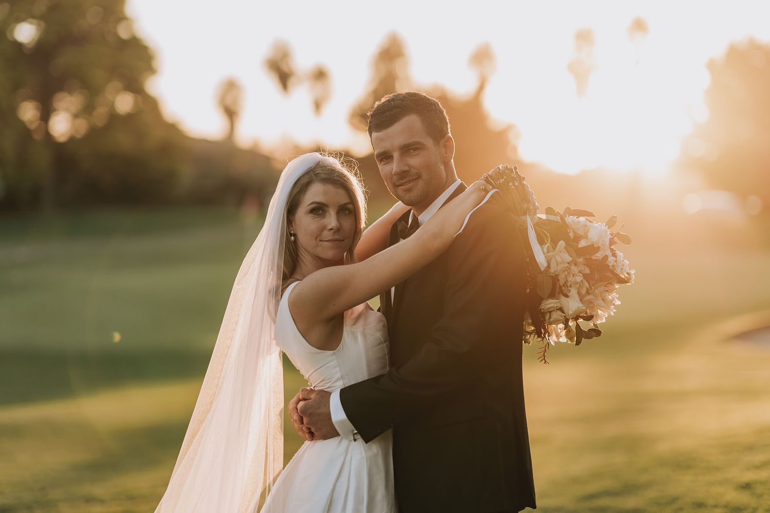 Newlyweds embrace in elegant wedding attire during golden hour sunset on a grassy lawn.
