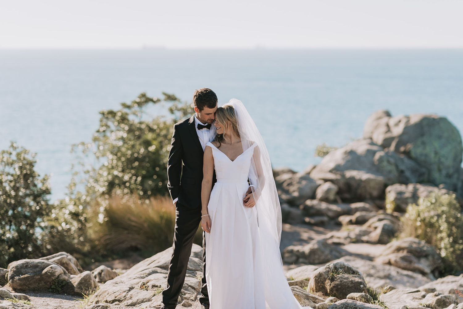 A couple in wedding attire embrace while standing on rocky terrain overlooking the ocean on a sunny day.