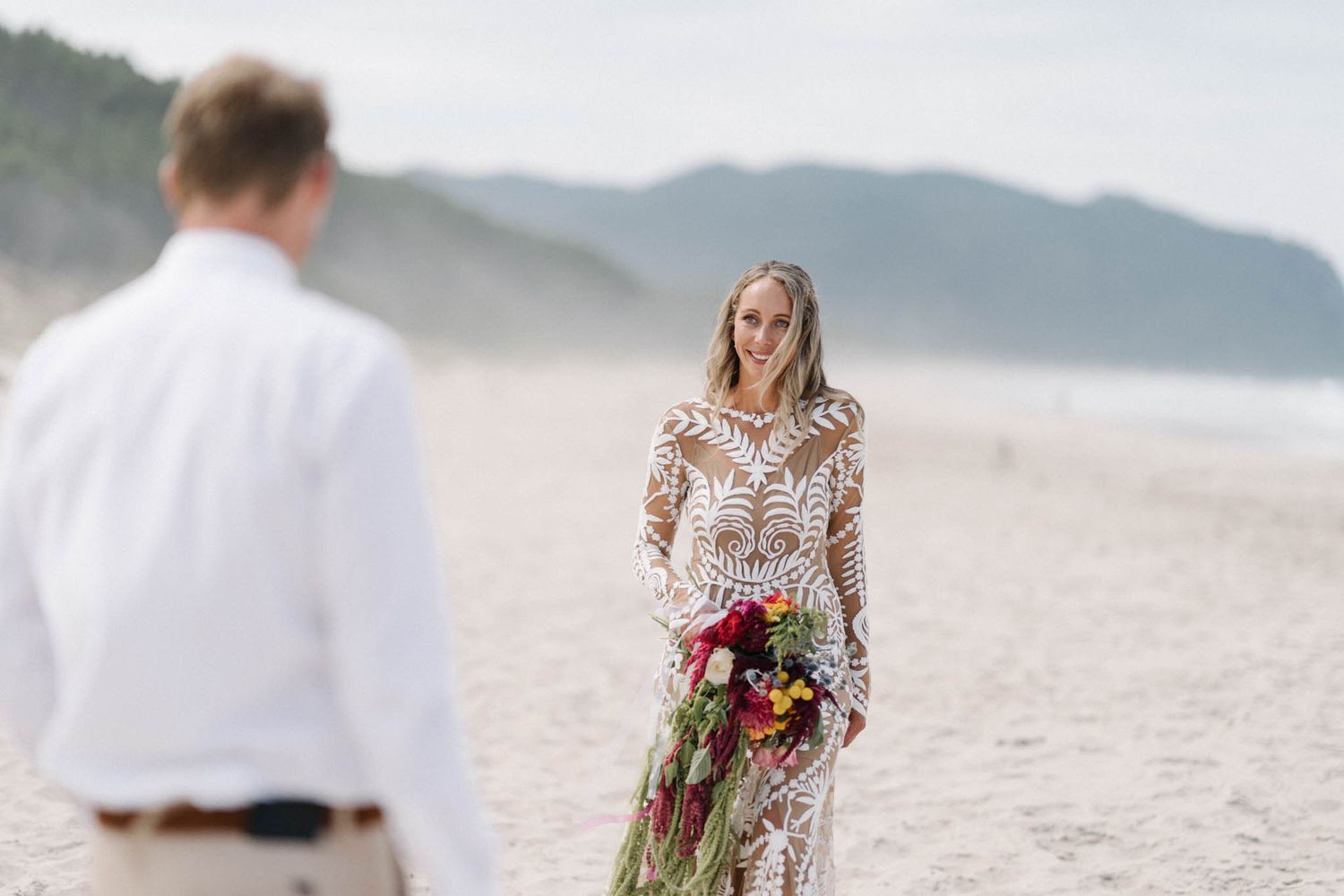 A couple having an intimate beach wedding celebration with mountain views and a lace dress on a misty shoreline.