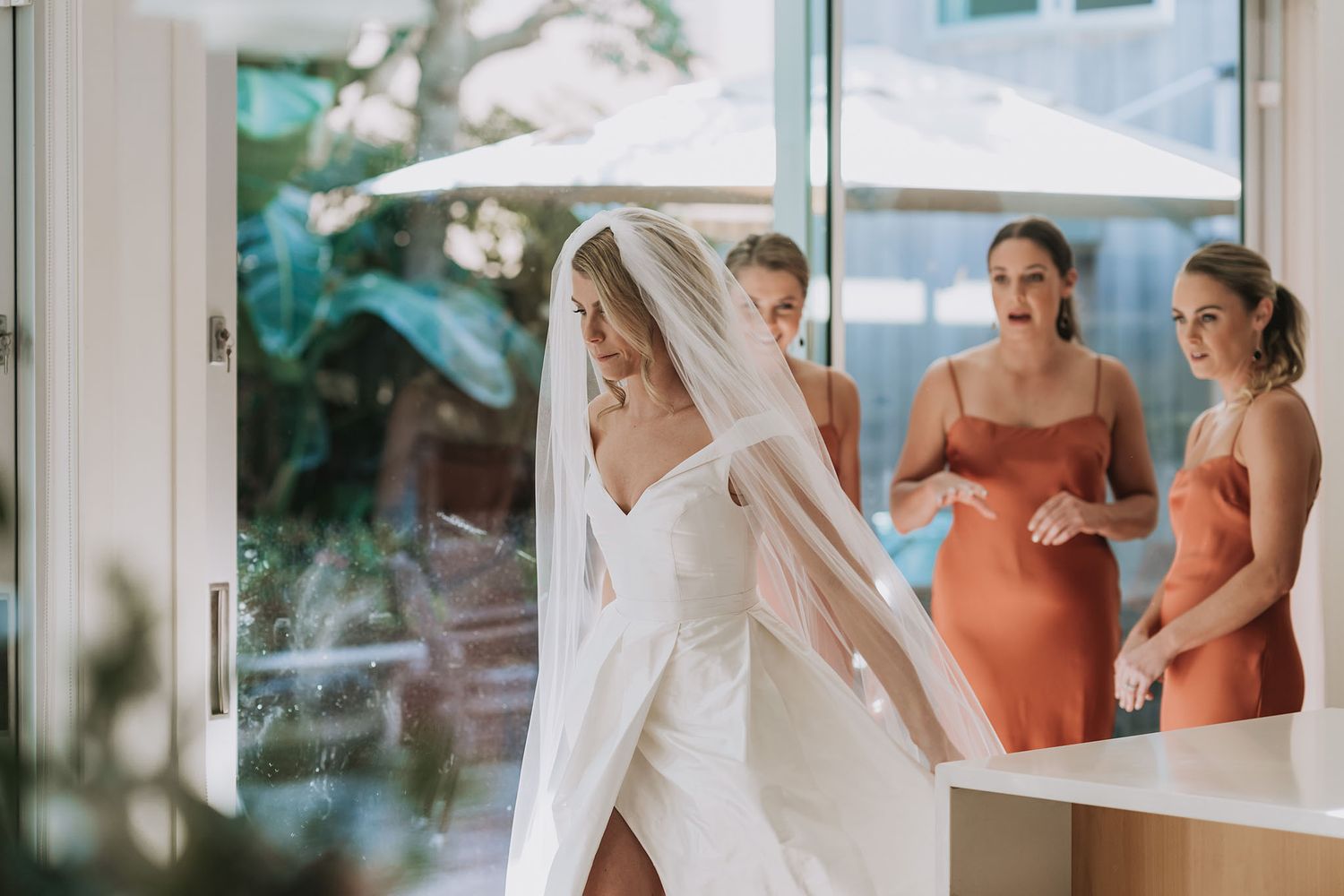 Bride in white wedding gown and veil getting ready in bright room with bridesmaids in coral dresses looking on.