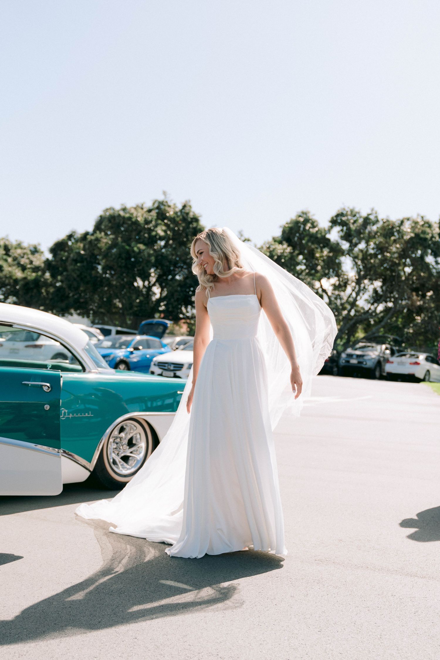Someone in a white wedding dress standing next to a vintage turquoise car in a sunny parking lot.
