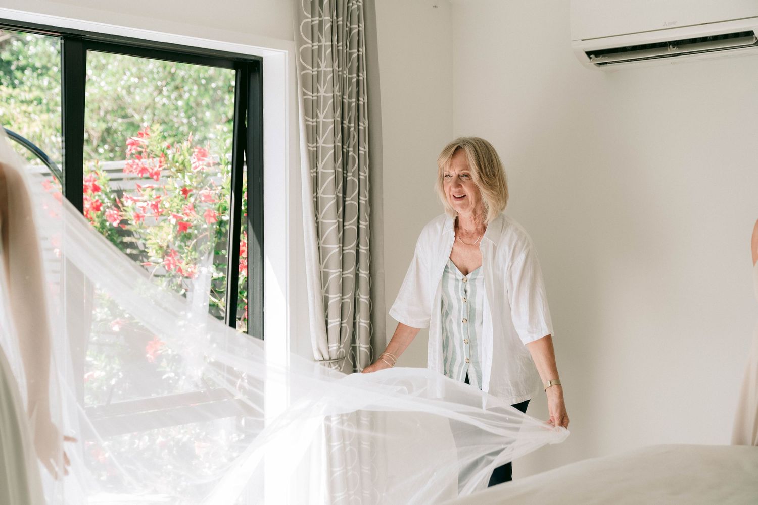 Someone adjusts white bedding or fabric in a bright room with large windows and flowing curtains.