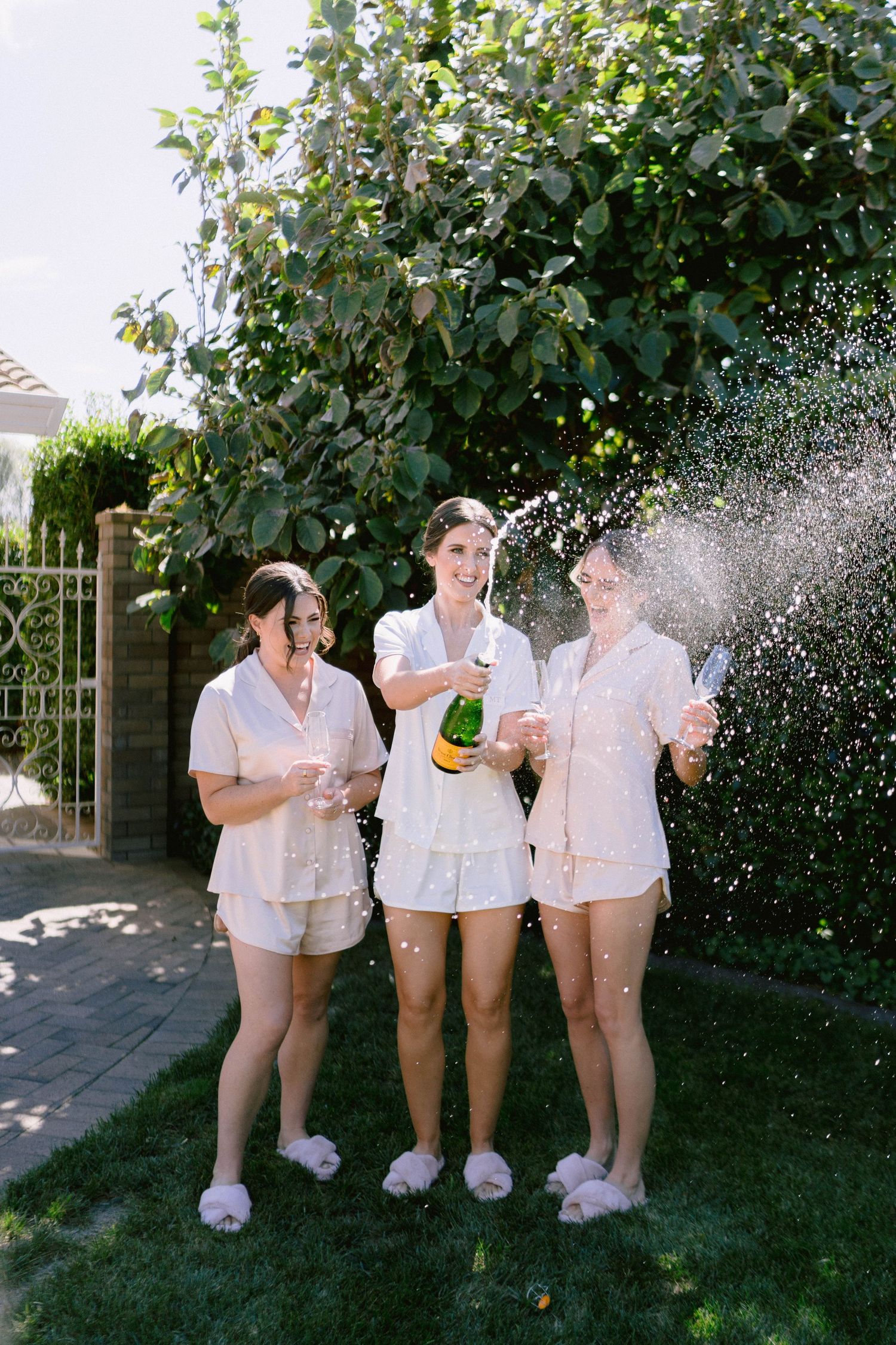 Three friends in matching white outfits celebrate with champagne in a sunny backyard garden setting.