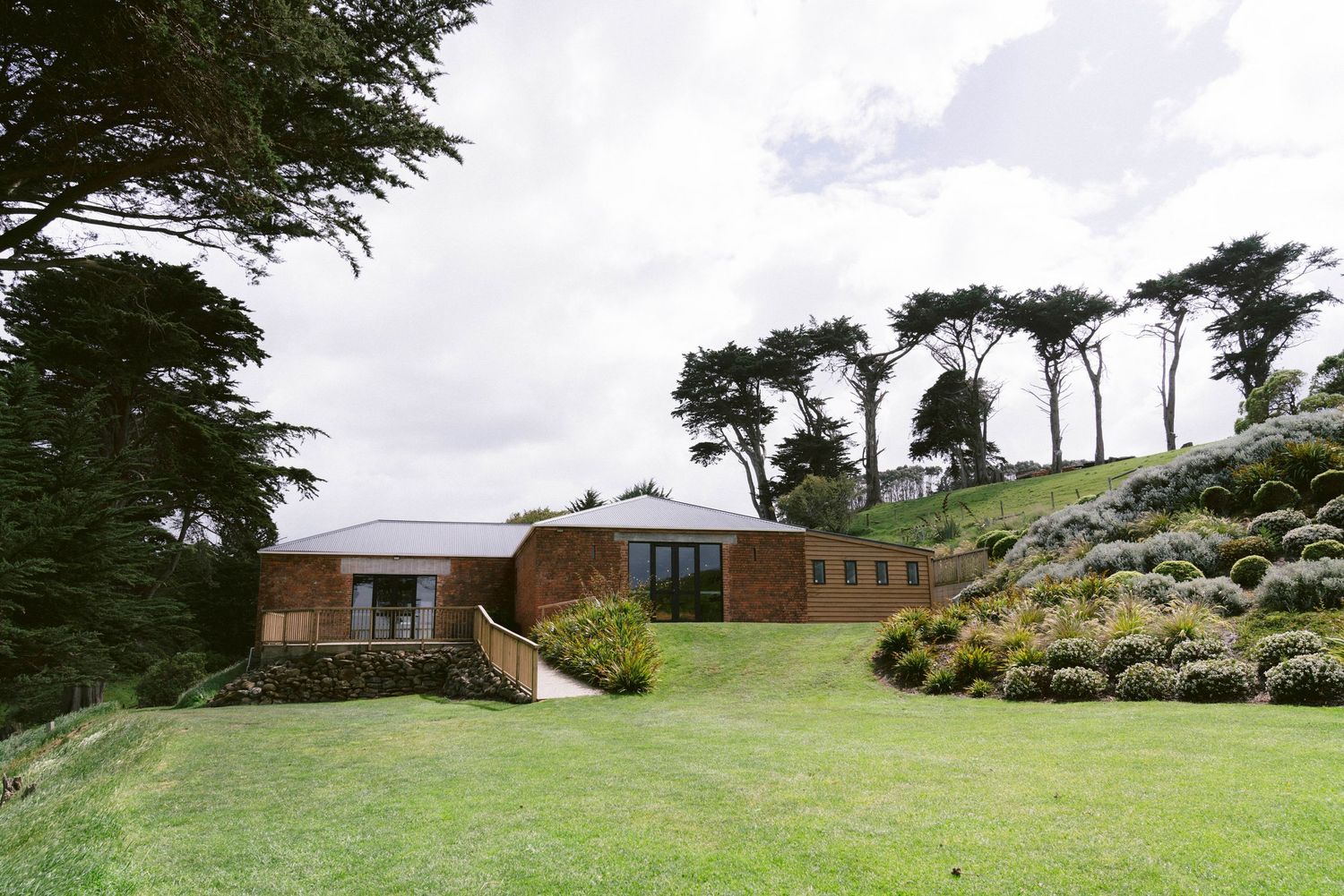 Modern brown brick building with glass windows nestled on a hillside surrounded by tall cypress trees and manicured lawn.