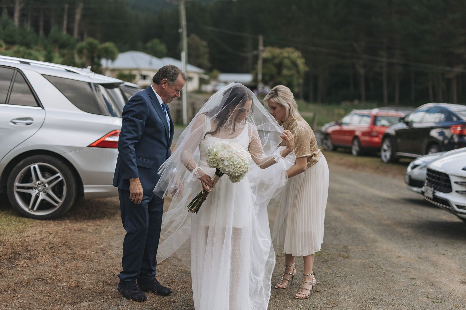 Wedding party gathered in a casual parking area with cars as a bride in white receives assistance with her dress and veil.