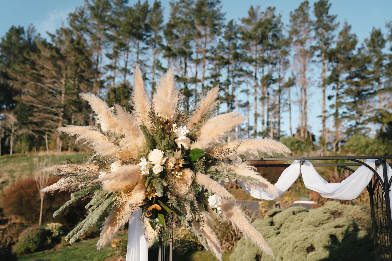 Rustic wedding arch decoration with pampas grass, dried flowers and white fabric against pine trees in natural setting.
